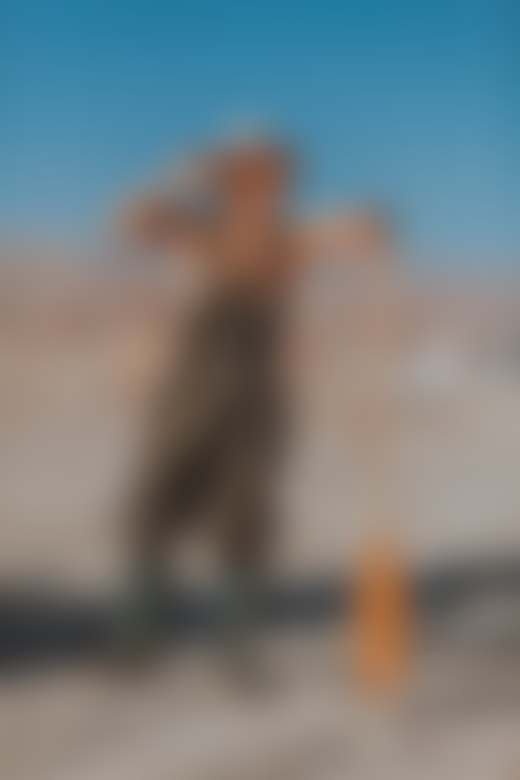 A person wearing overalls, a hat, and boots leans against a paddle while standing on the shore of the Salton sea.