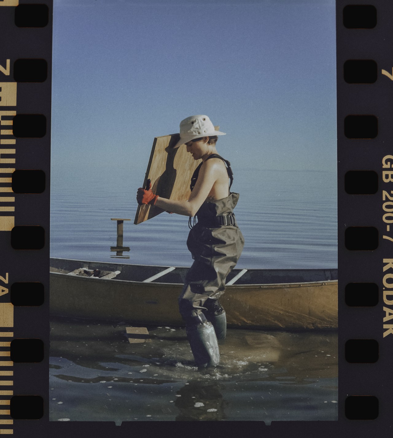 A person wearing overalls, a hat, and boots lifts a piece of wood into a canoe on the shore of the Salton sea