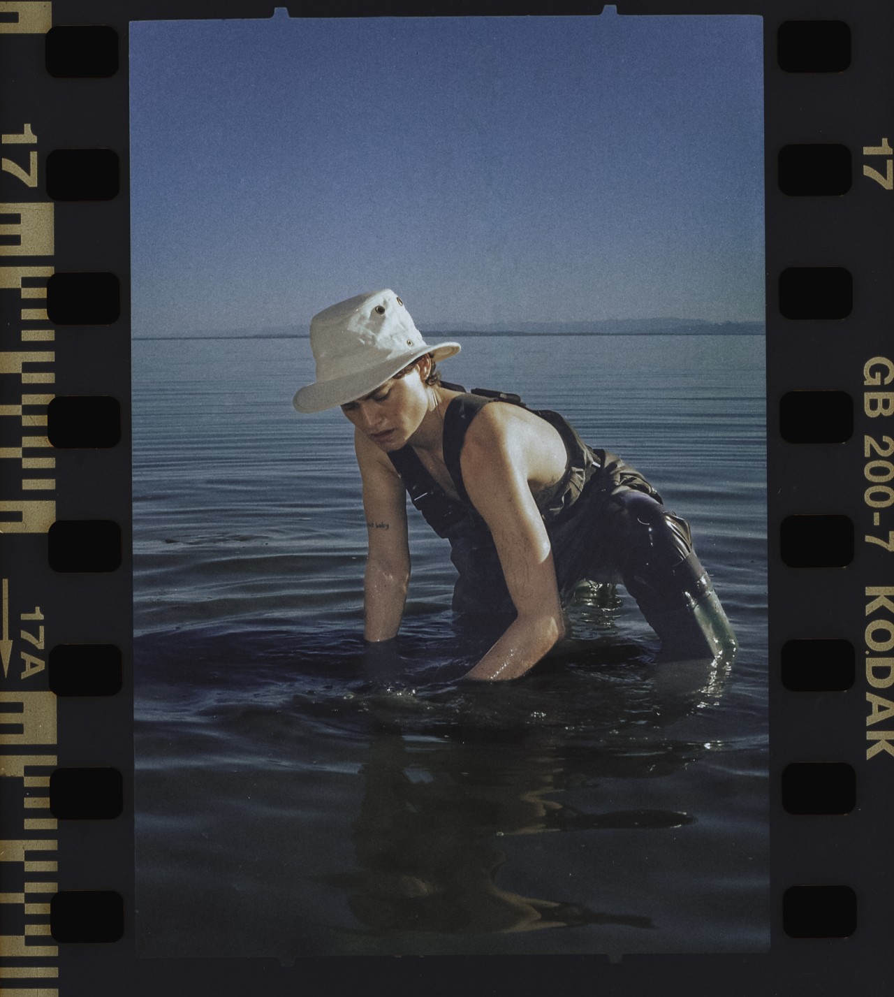A person wearing overalls, a hat, and boots kneels in the Salton sea.