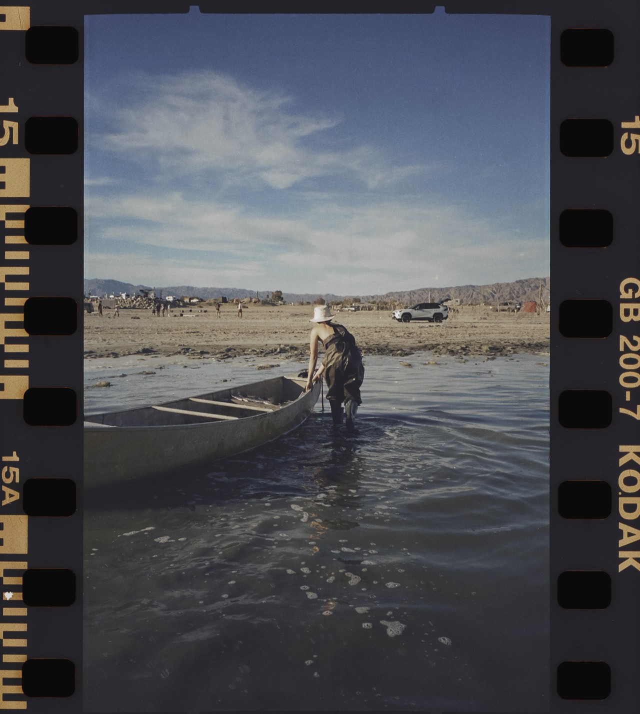 A person wearing overalls, a hat, and boots pulls a canoe onto the shore of the Salton sea.