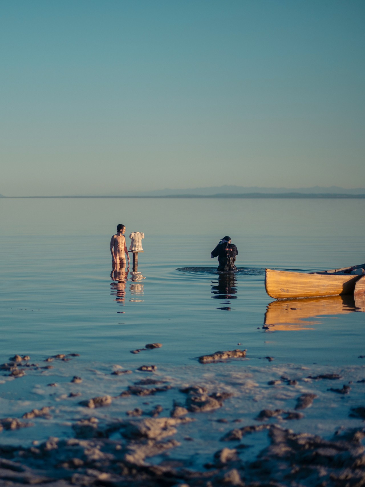 A person stands in the shallow water of the Salton Sea next to a sculpted torso of a human. Another person, standing next to a canoe in the water, photographs them.