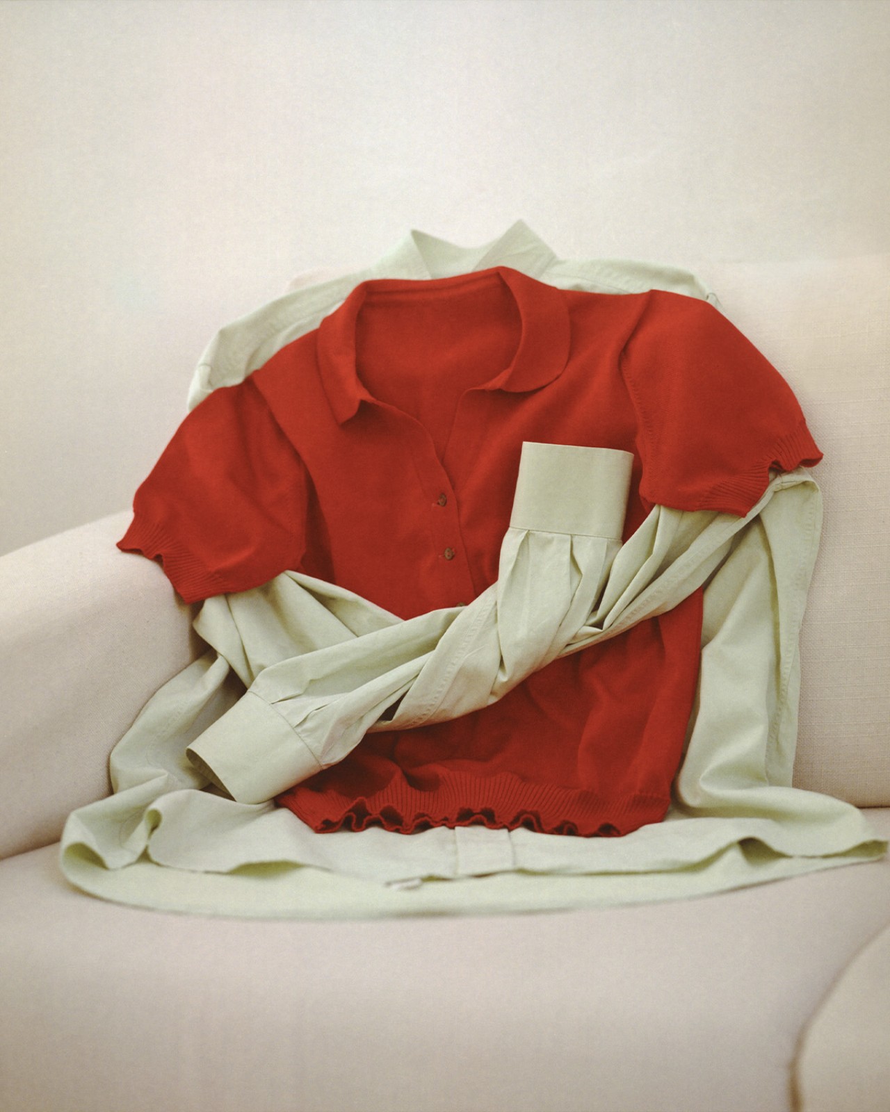 A red polo shirt is draped over a light green button-up shirt on a beige couch.