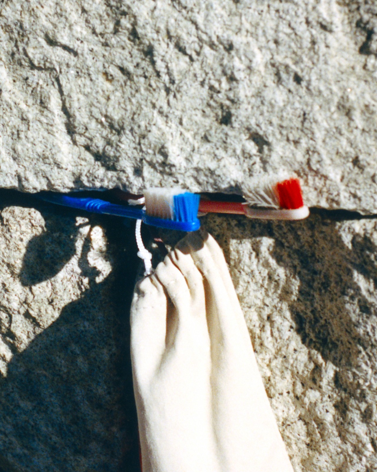 A red and blue toothbrush are attached to the side of a rock with a small white cloth bag hanging from them both.