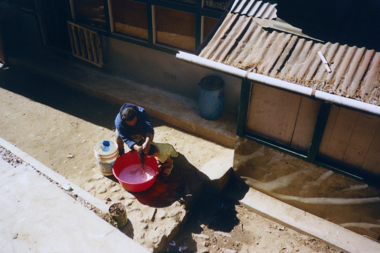 A person washes their belonging in a red soapy bucket outside their home.