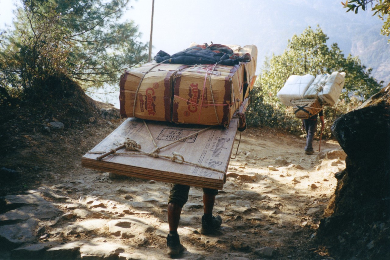 Two people carry large packages on their backs in the Himalaya hills.