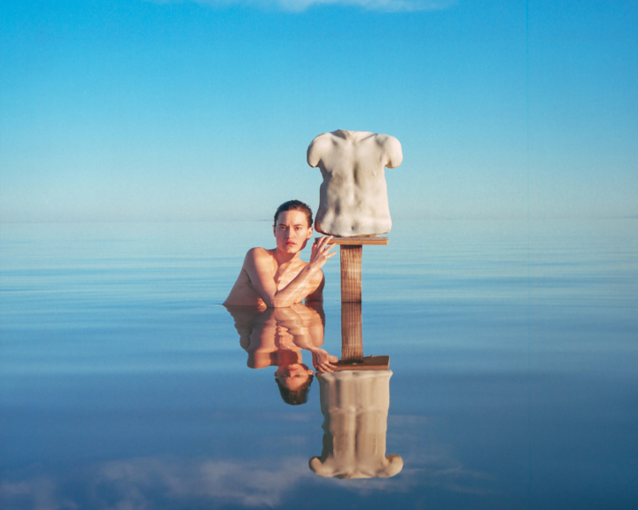 A person sits in the shallow water of the Salton Sea next to a sculpted torso of a human.