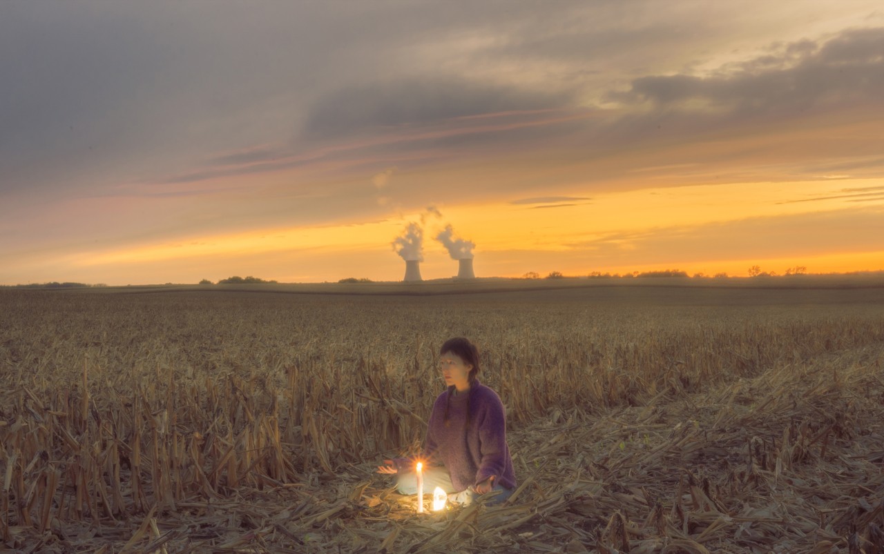 A person sits in a grassy field with a burning candle in front of them.