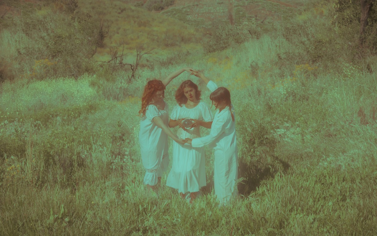 Two girls connect their arms to make a circle around another girl. They are dressed in white gowns and standing in a grass field.