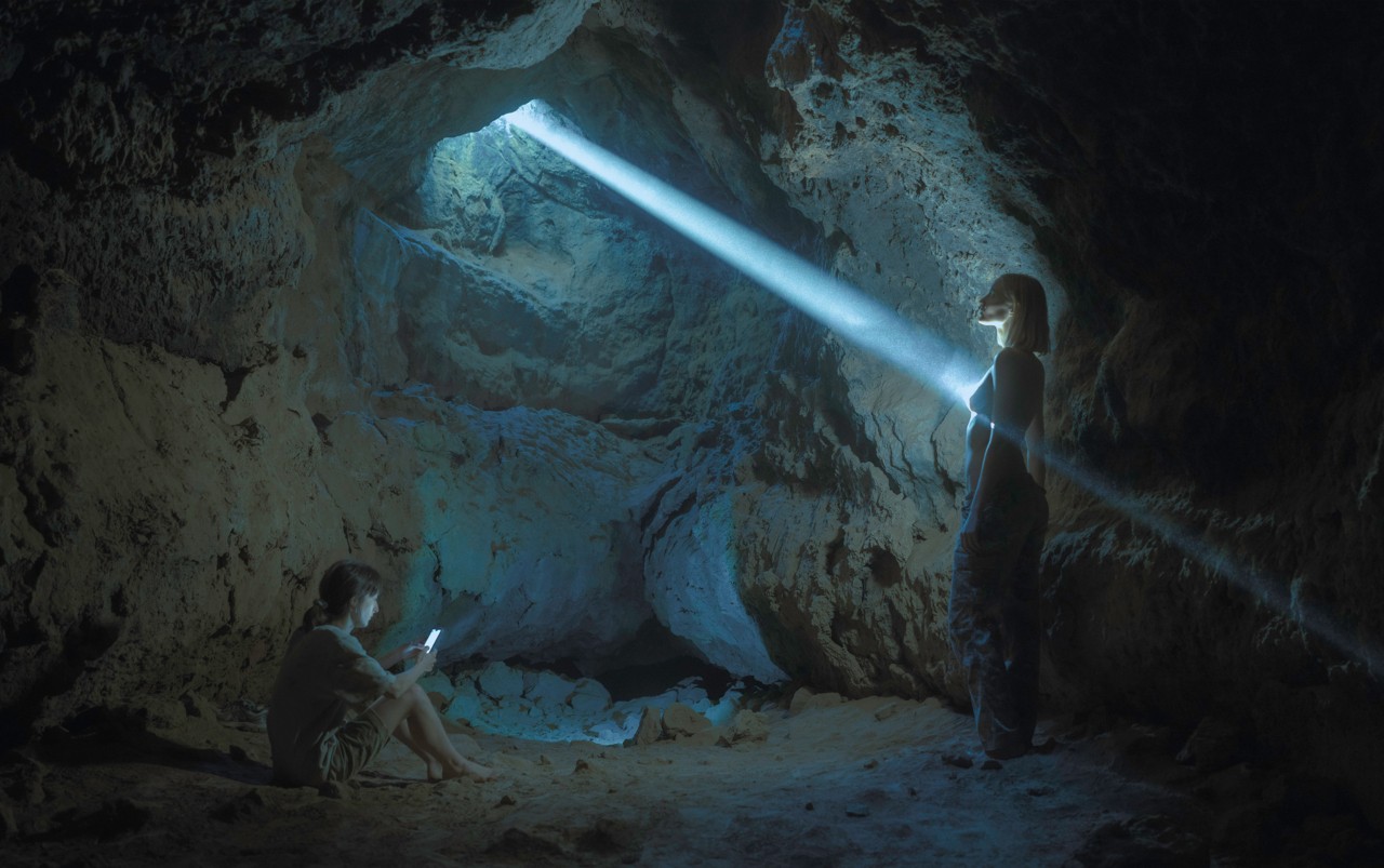Two people are inside a cave. One sits down while looking at their phone and the other stands toward an opening at the top of the cave, where light pierces through.