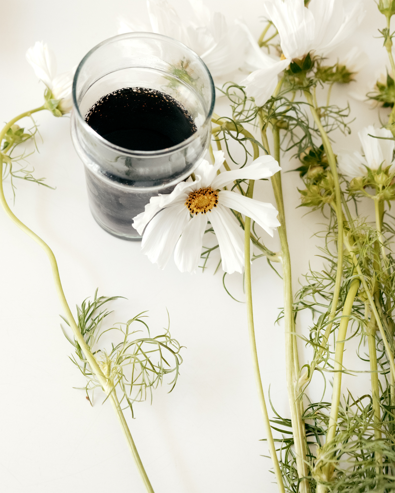 A glass cup filled with soda sits on a white table surrounded by white flowers.