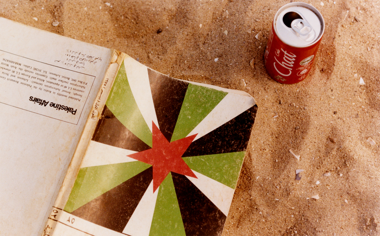 A book called Palestine Affairs with a cover using the Palestinian flag colors lays in the sand next to a can of Gaza cola.