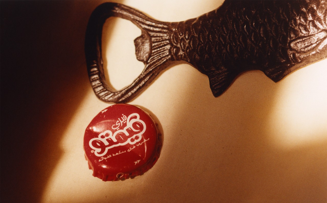 A black can opener lays on the table next to a Gaza cola bottle cap.
