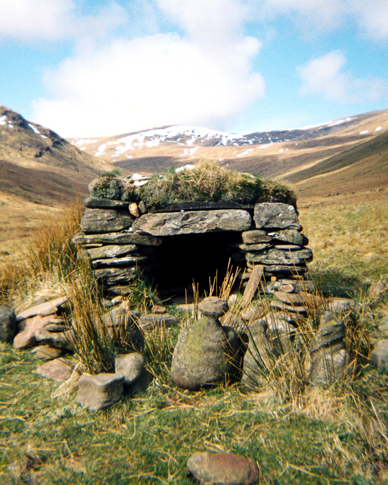 A shelter made from stacked rocks sits on a plain with snow capped mountains in the distance.