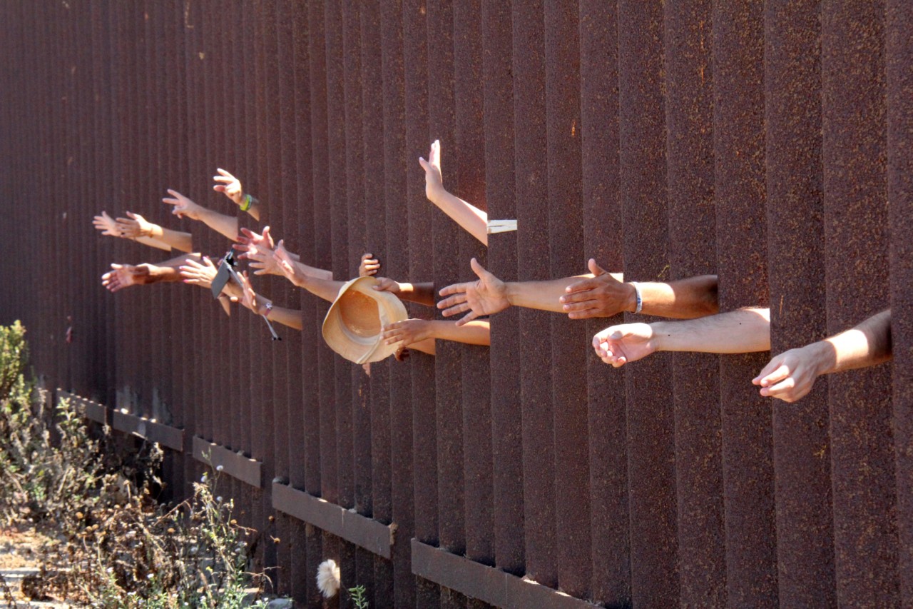Several hands reach through metal poles separating Tijuana from San Diego.