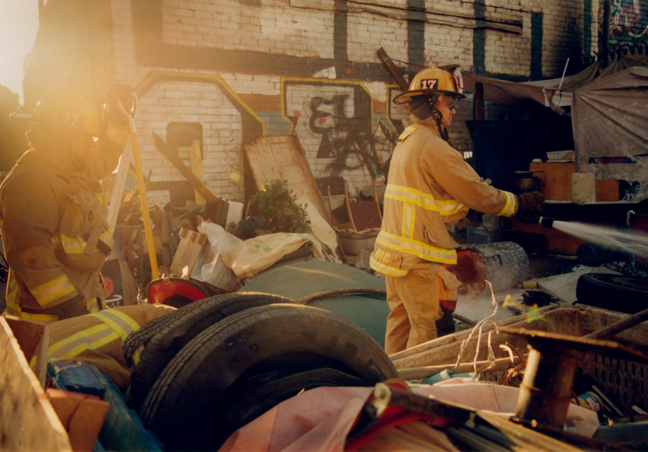 Two firefighters walk through rubble near a house.
