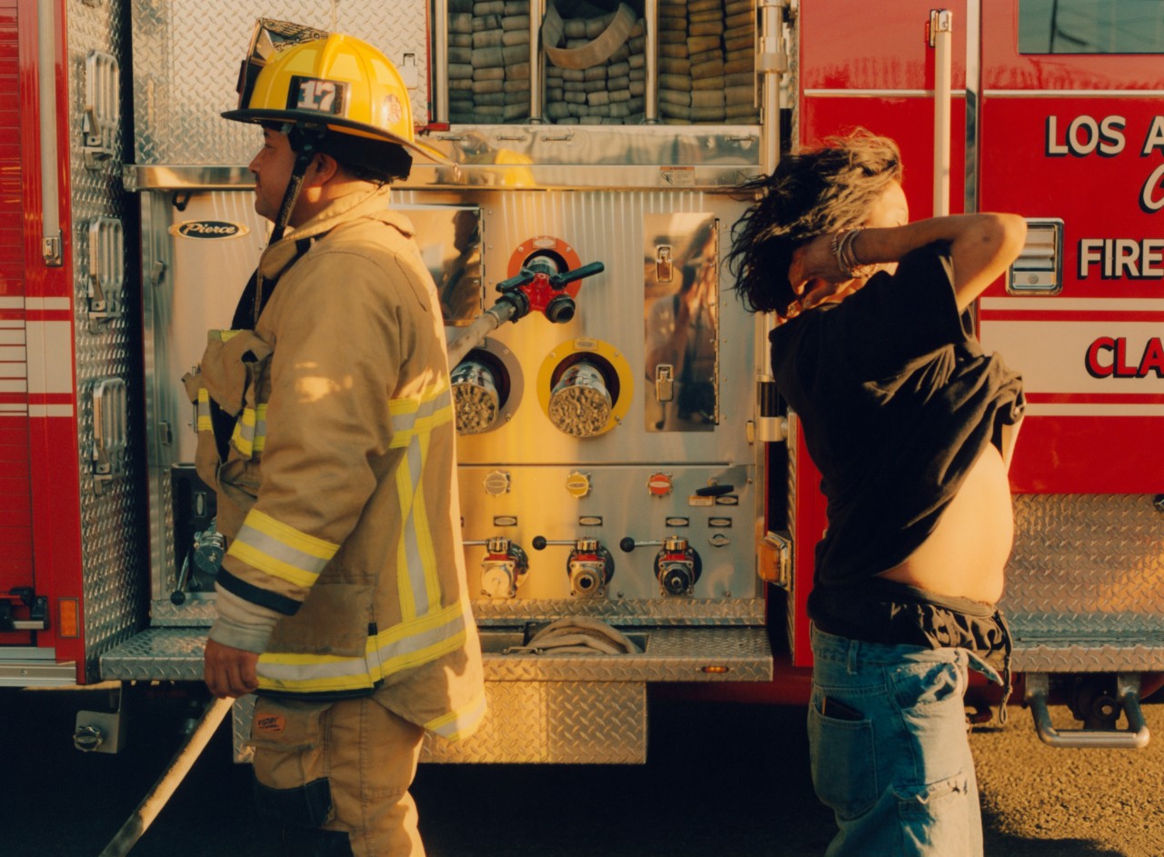 Two Southern California firefighters stand facing away from one another in front of a fire truck.