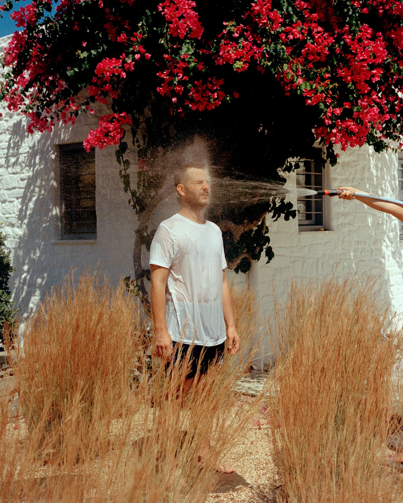 A man stands among dry grass in front of a white house getting sprayed in the face with a hose.