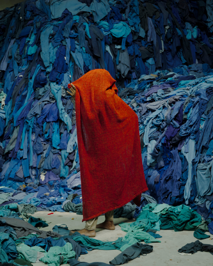 A person wearing a red drape covering their body and face stands in front of a large pile of blue cloths.
