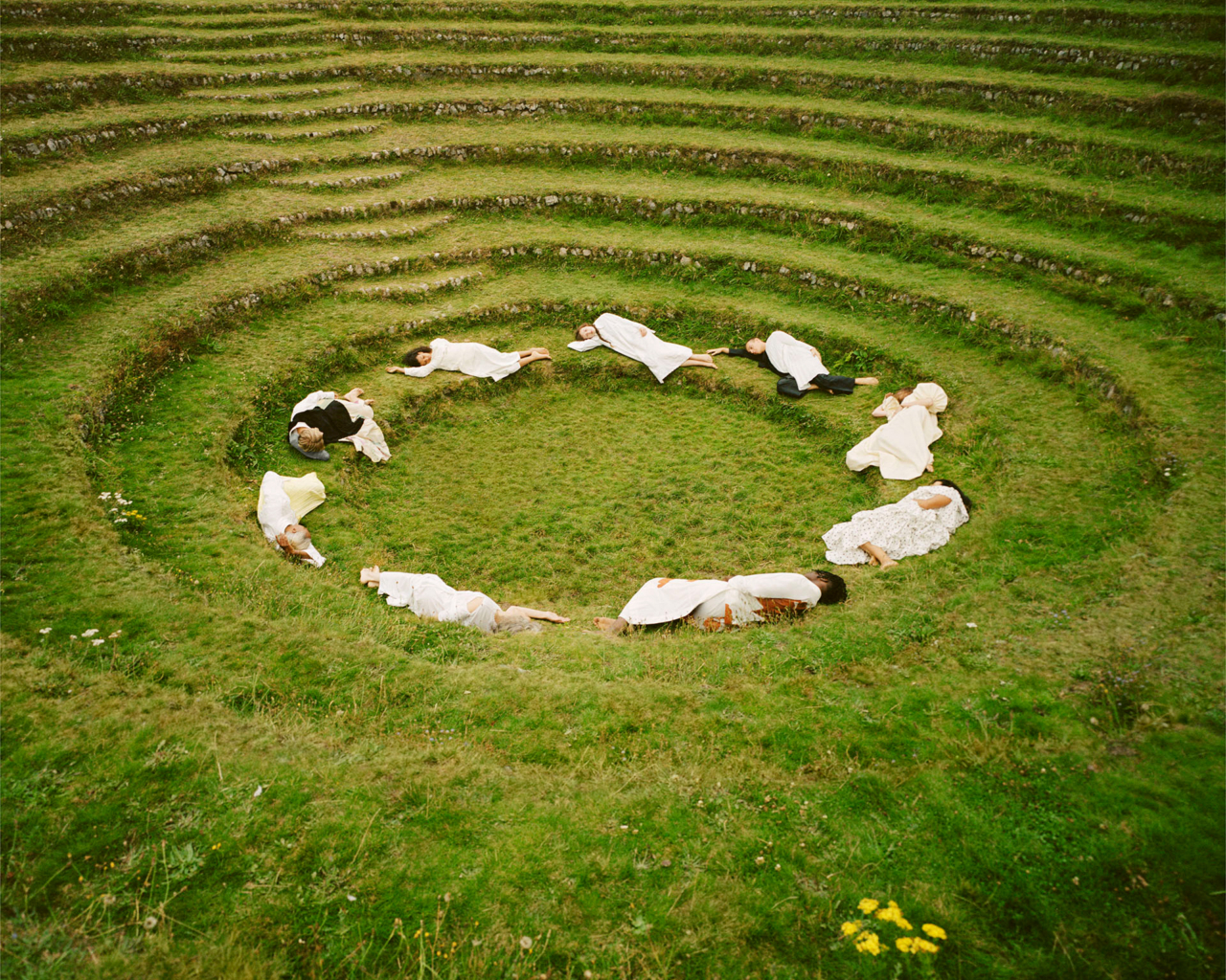 A group of people lie down in a circle on a green field.