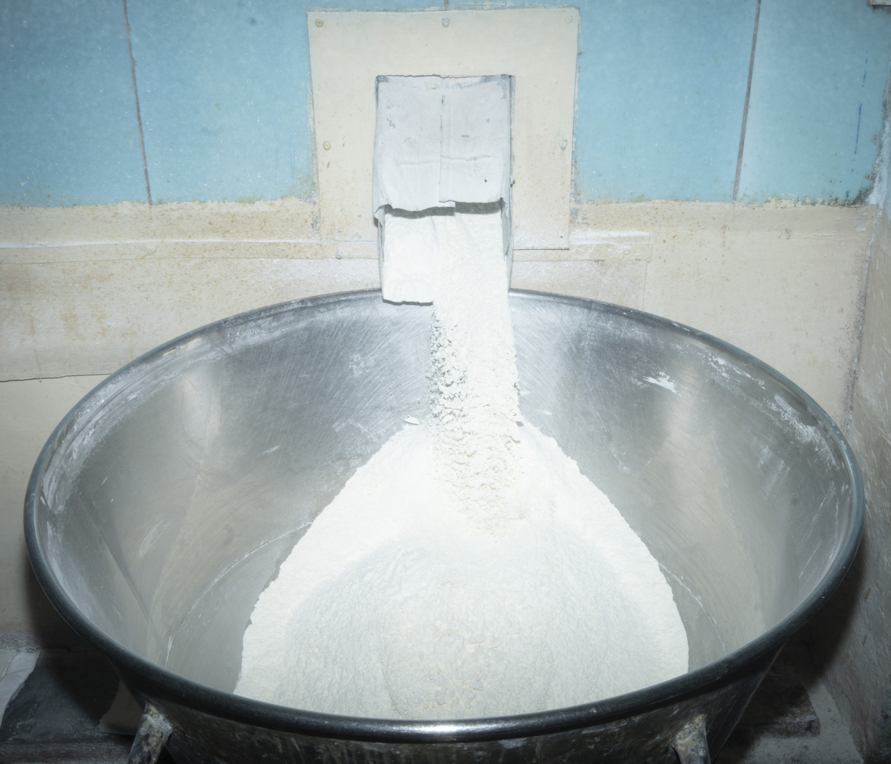 A white powder pours from a square spout into a large silver container.
