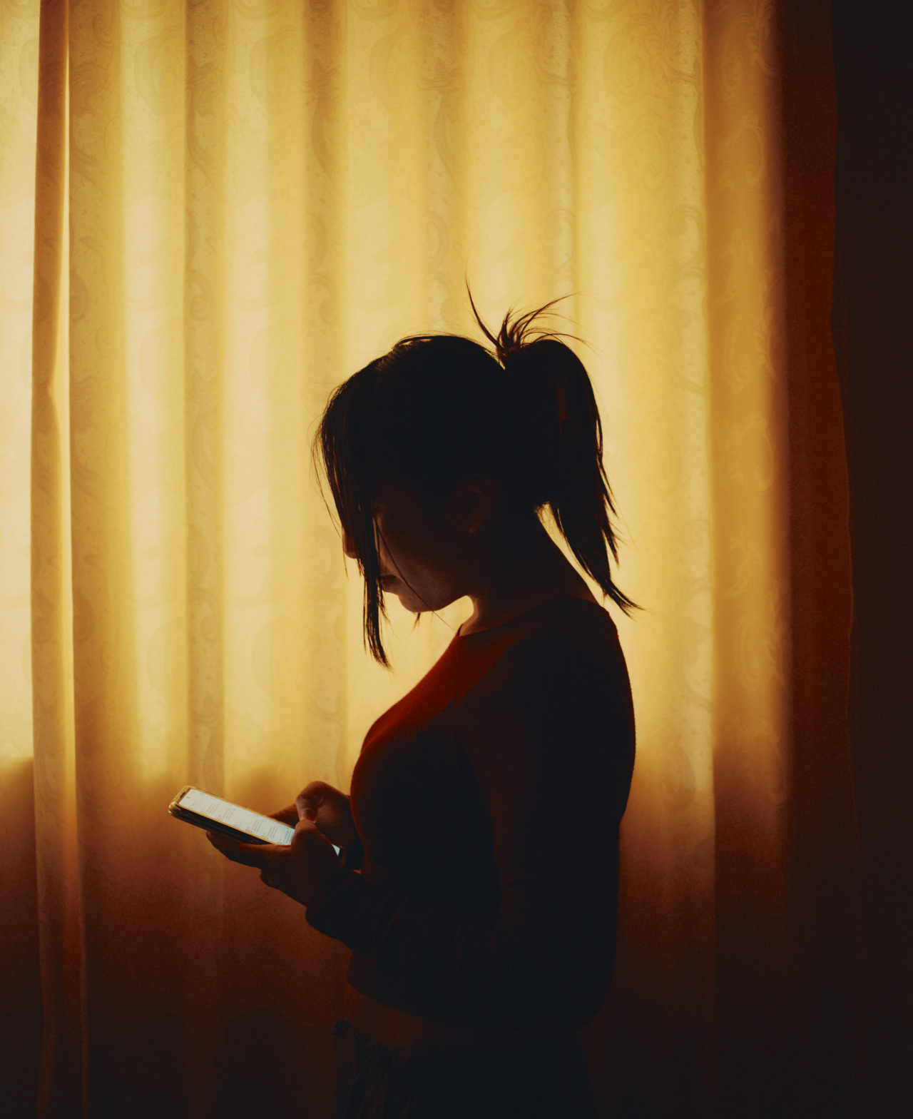 A teenagers looks at their phone while standing in front of a window.