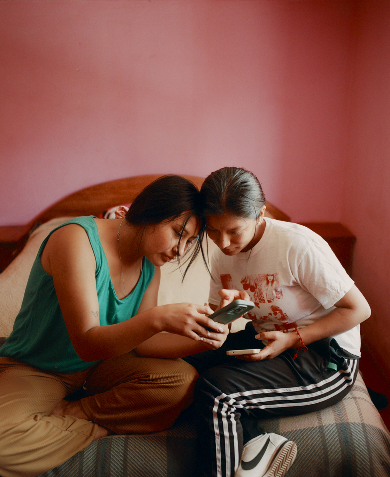 Two teens sit on a bed looking at a phone.