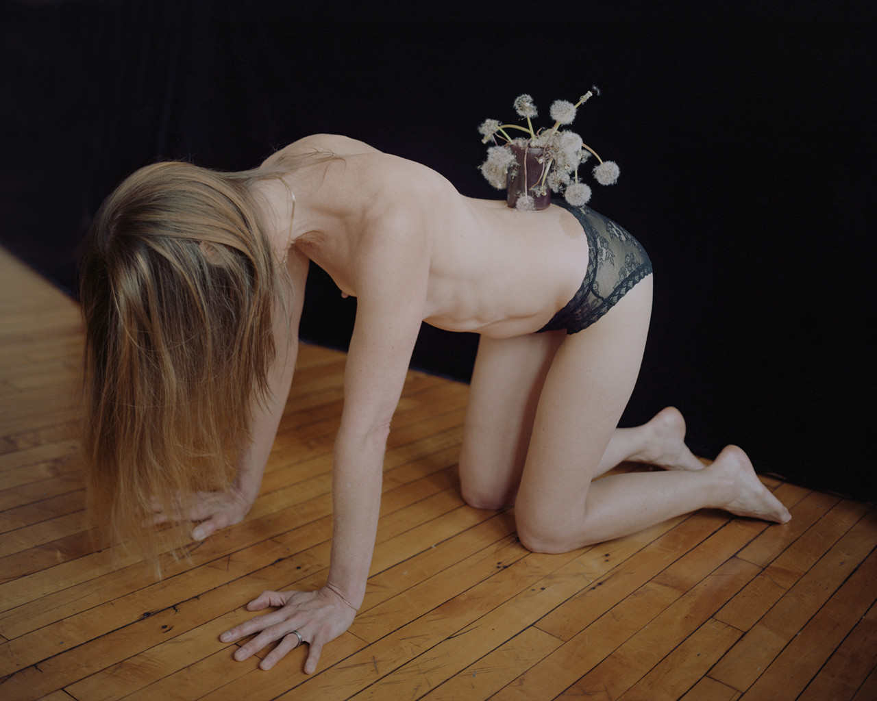 A half nude woman balances a flower pot of dandelions on her lower back.
