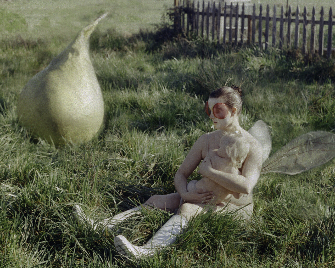 A mother and child dressed as fruit flies sit in the grass with a large pear sculpture in the background.