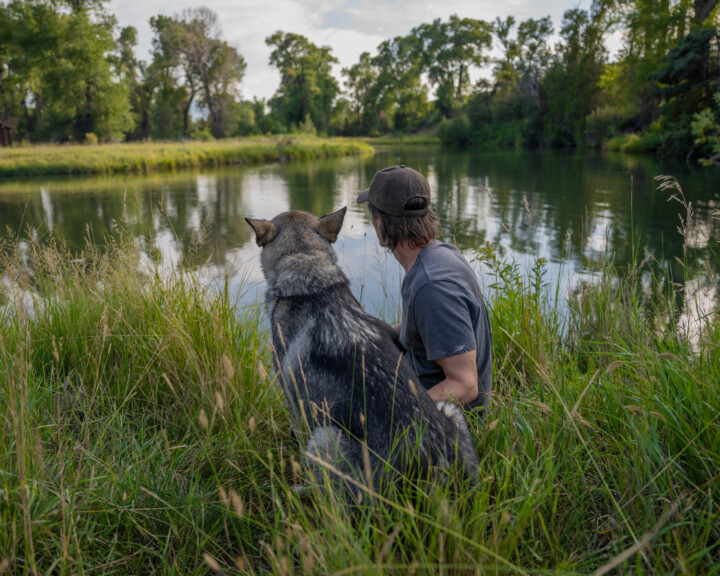A man sits in front of a lake with a wolf dog beside him.